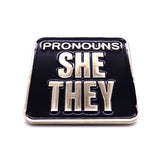 Picture shows backside of pronoun badge - "PRONOUNS / SHE/ THEY" silver metal and black enamel
